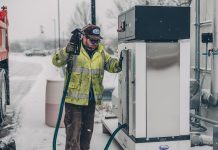 A man wearing a jacket refuels a truck at a gas station during winter with snow falling.