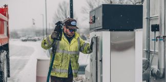 A man wearing a jacket refuels a truck at a gas station during winter with snow falling.