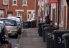 A street scene in a residential area with garbage bins and parked cars along the sidewalk.