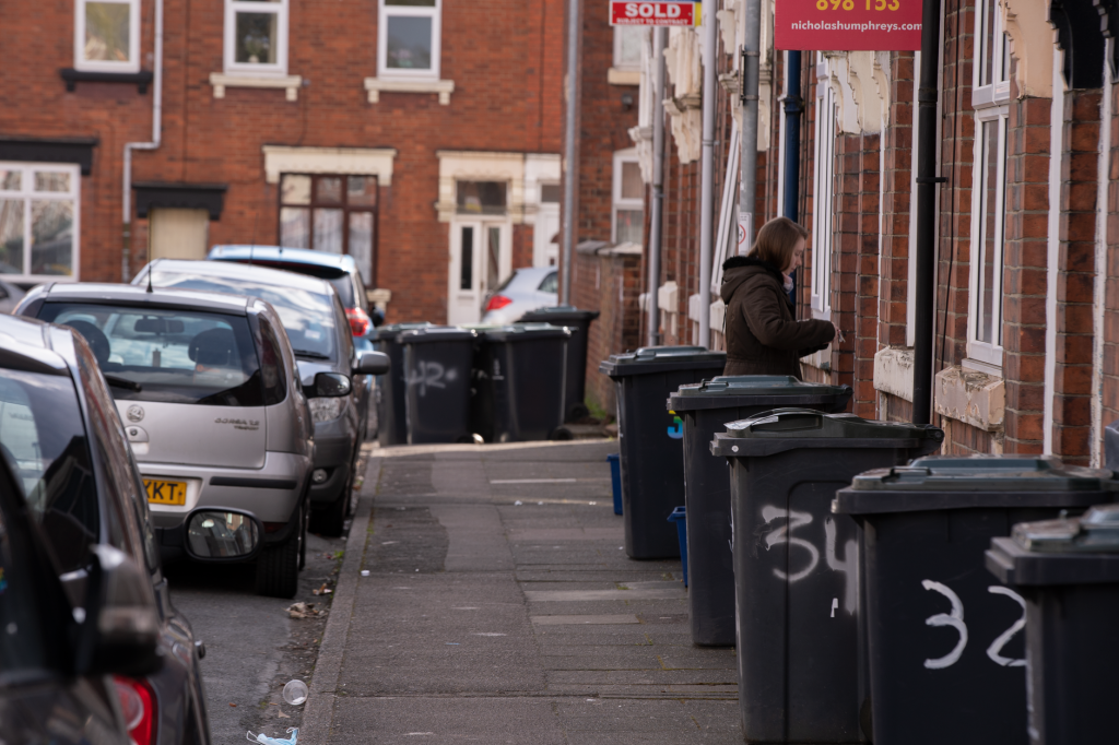 A street scene in a residential area with garbage bins and parked cars along the sidewalk.