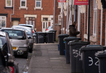 A street scene in a residential area with garbage bins and parked cars along the sidewalk.
