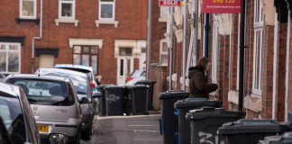 A street scene in a residential area with garbage bins and parked cars along the sidewalk.