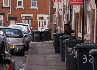 A street scene in a residential area with garbage bins and parked cars along the sidewalk.