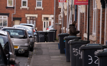 A street scene in a residential area with garbage bins and parked cars along the sidewalk.