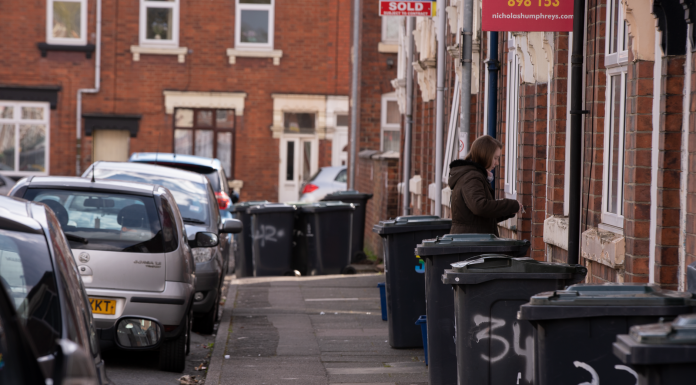 A street scene in a residential area with garbage bins and parked cars along the sidewalk.