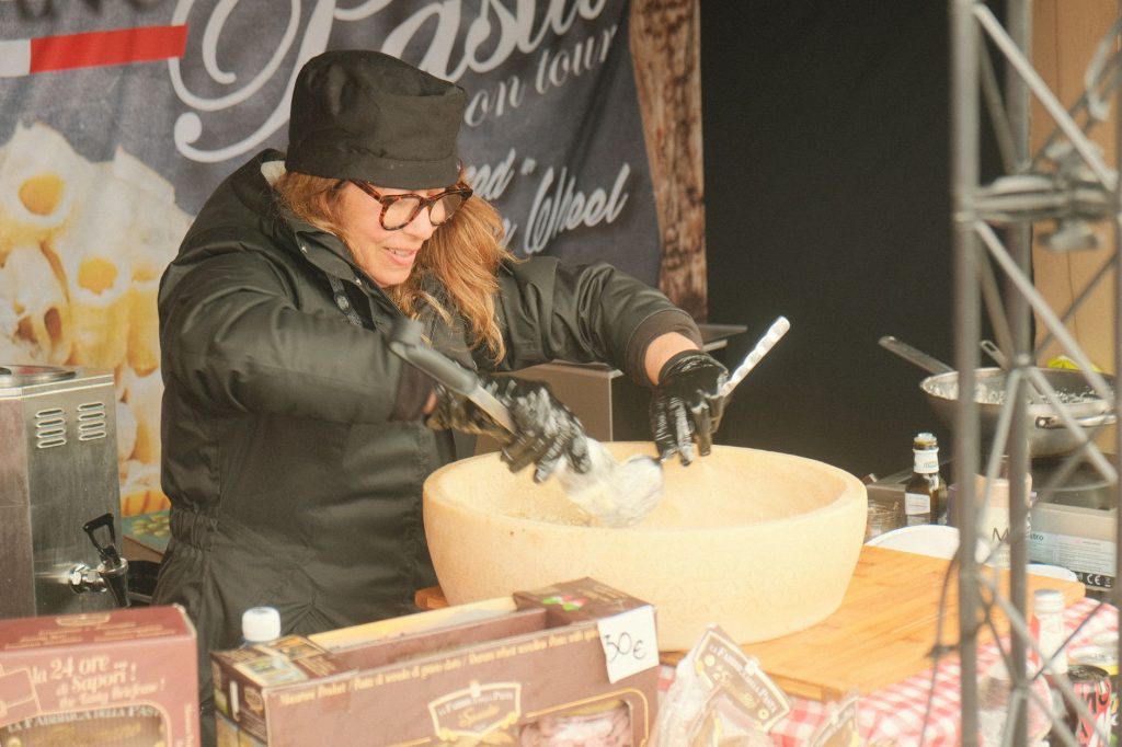 Female chef preparing pasta in a large parmesan wheel at an outdoor food stall, showcasing culinary techniques.