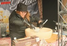 Female chef preparing pasta in a large parmesan wheel at an outdoor food stall, showcasing culinary techniques.