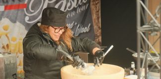Female chef preparing pasta in a large parmesan wheel at an outdoor food stall, showcasing culinary techniques.