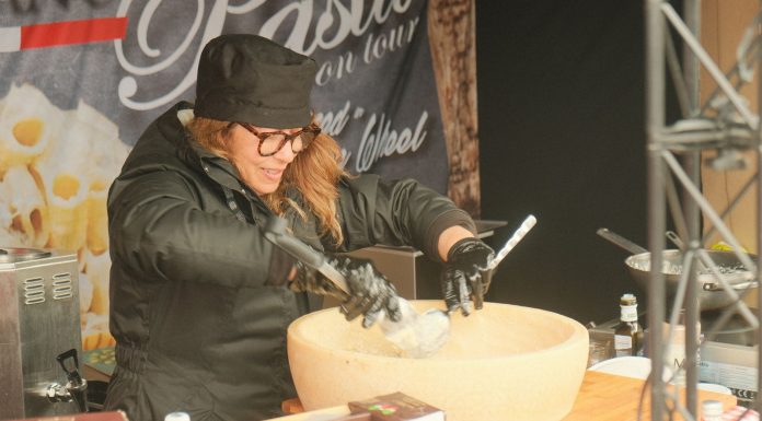 Female chef preparing pasta in a large parmesan wheel at an outdoor food stall, showcasing culinary techniques.