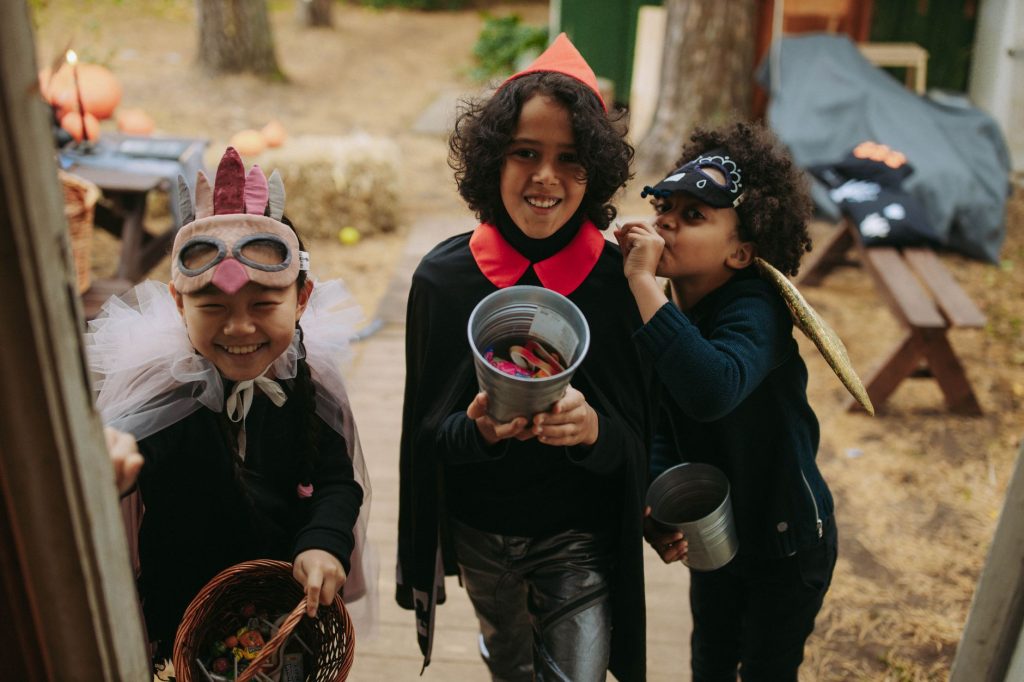 Three children in Halloween costumes joyfully trick-or-treating with candy buckets.