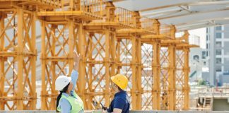 Two engineers in hardhats discuss projects at a busy construction site.