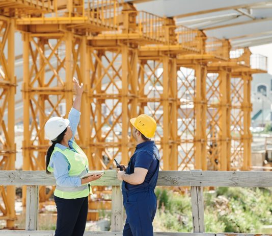 Two engineers in hardhats discuss projects at a busy construction site.
