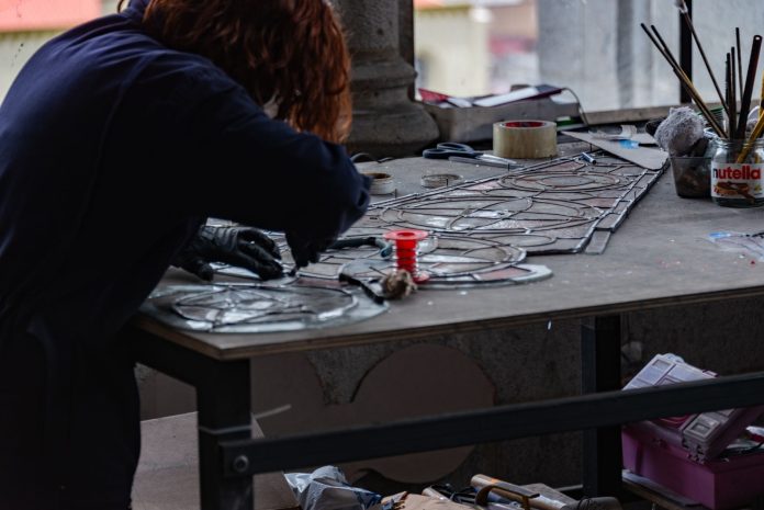 ltima-hora-nuevo-estudio-de-vitrales-abre-en-jers Redhead artisan working meticulously on stained glass artwork at a workshop table.