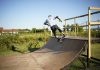 Skateboarder executing a trick on a wooden outdoor skate ramp with lush greenery in the background.